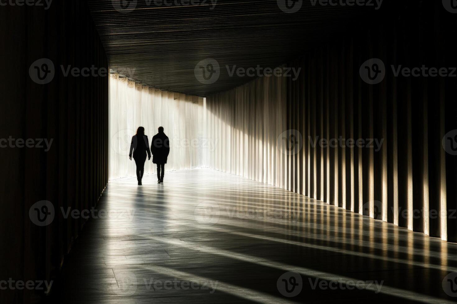 Two people walking down a long hallway with light coming from the end photo