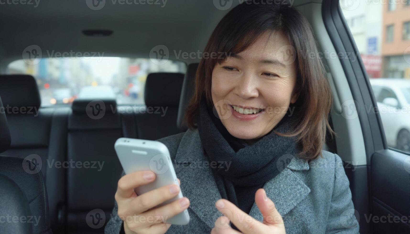 A middle aged Asian woman with shoulder length black hair, wearing a gray coat and scarf, smiles while using a smartphone in a car photo