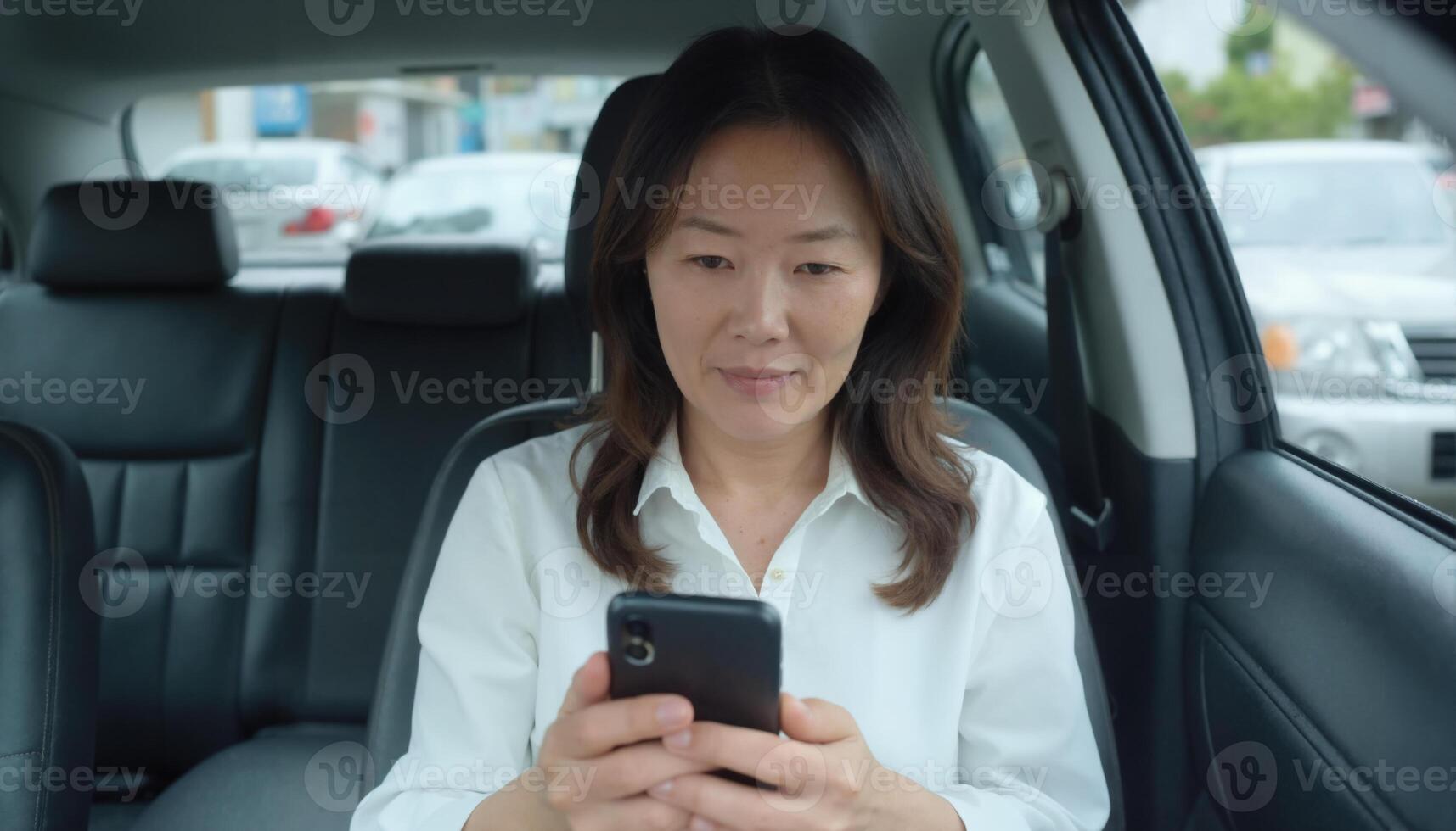 Asian woman in white shirt, sitting in car, using smartphone, with city background, looking focused photo