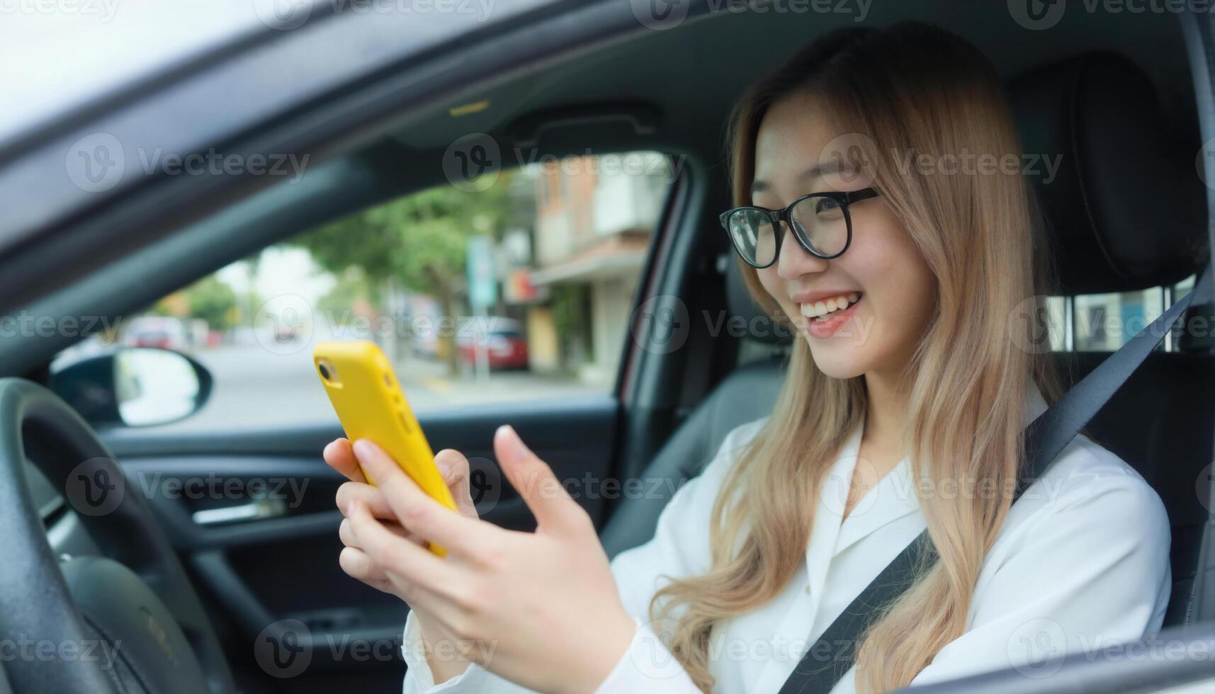 A smiling Asian woman with glasses, in a white shirt, uses a yellow phone in a car on a sunny day photo