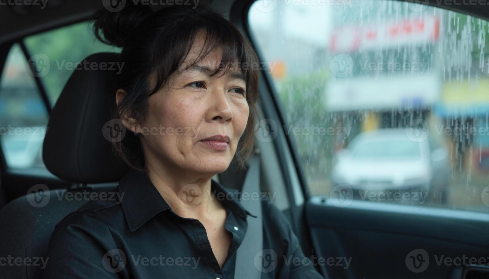 A middle aged Asian woman with short black hair and bangs sits in a car, looking out a rain speckled window photo