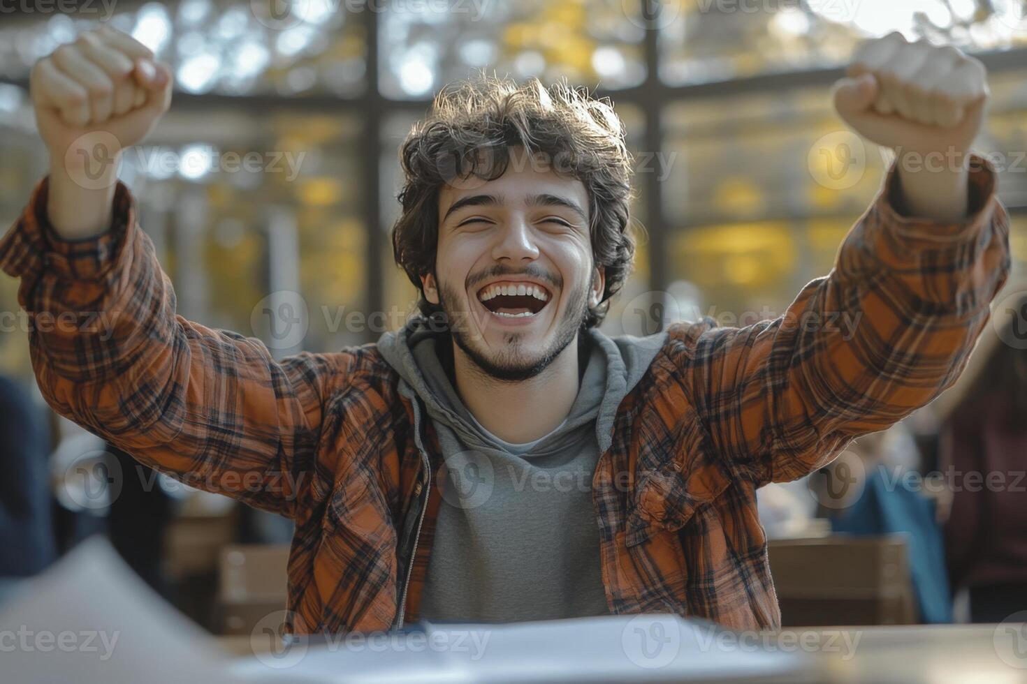 A young man with curly hair sits at a table, arms raised in joyful celebration photo
