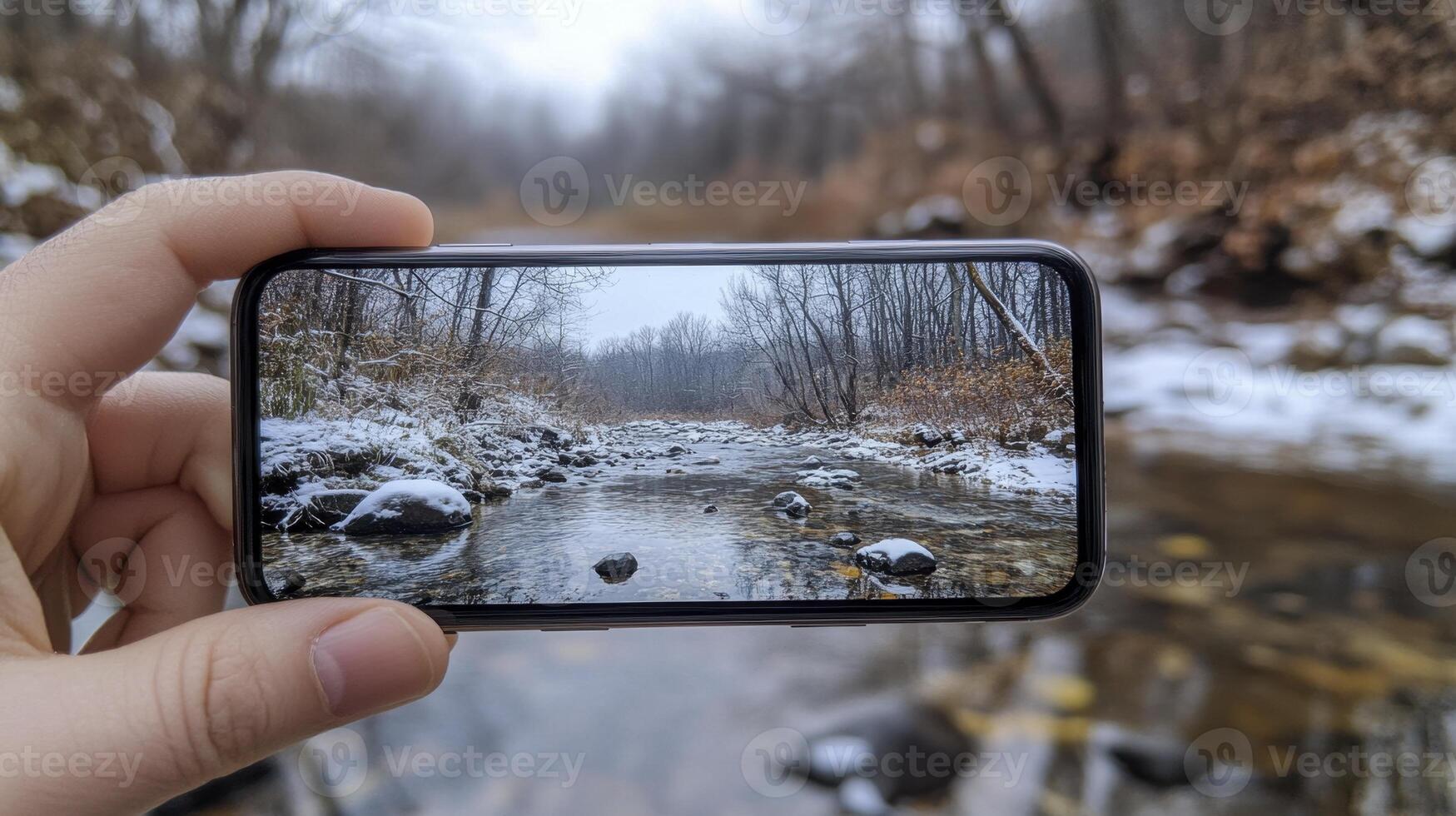 A hand holds a smartphone capturing a winter stream scene. Snow covers the banks, trees are bare, and the water is calm photo