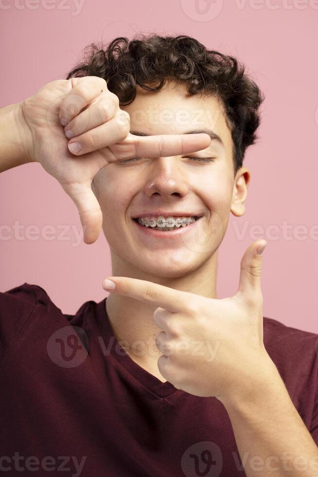 Portrait of excited, smiling teenager, boy with braces showing frame from fingers on pink background photo