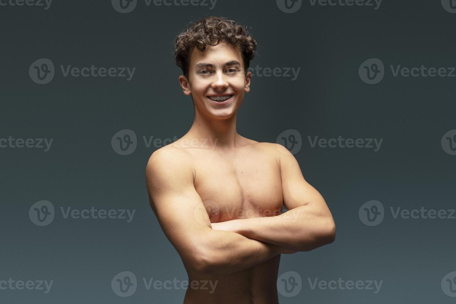 Portrait of attractive teen boy with bare torso, crossed arms posing isolated on gray background photo