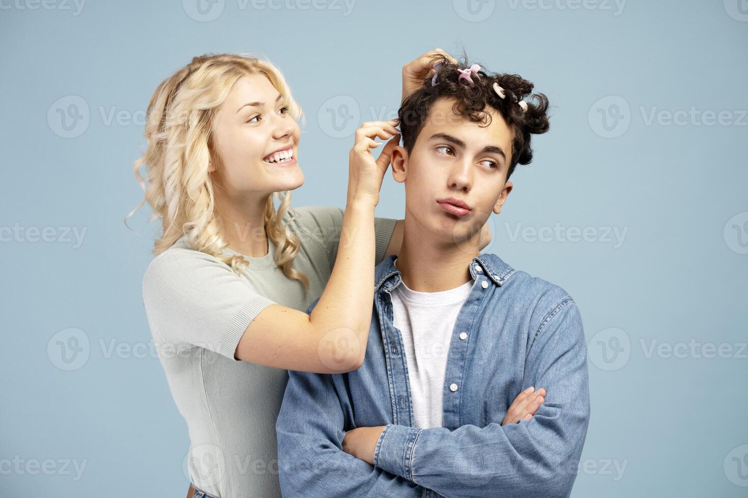 Happy, attractive couple, smiling girl doing hairstyle to handsome boy isolated on blue background photo