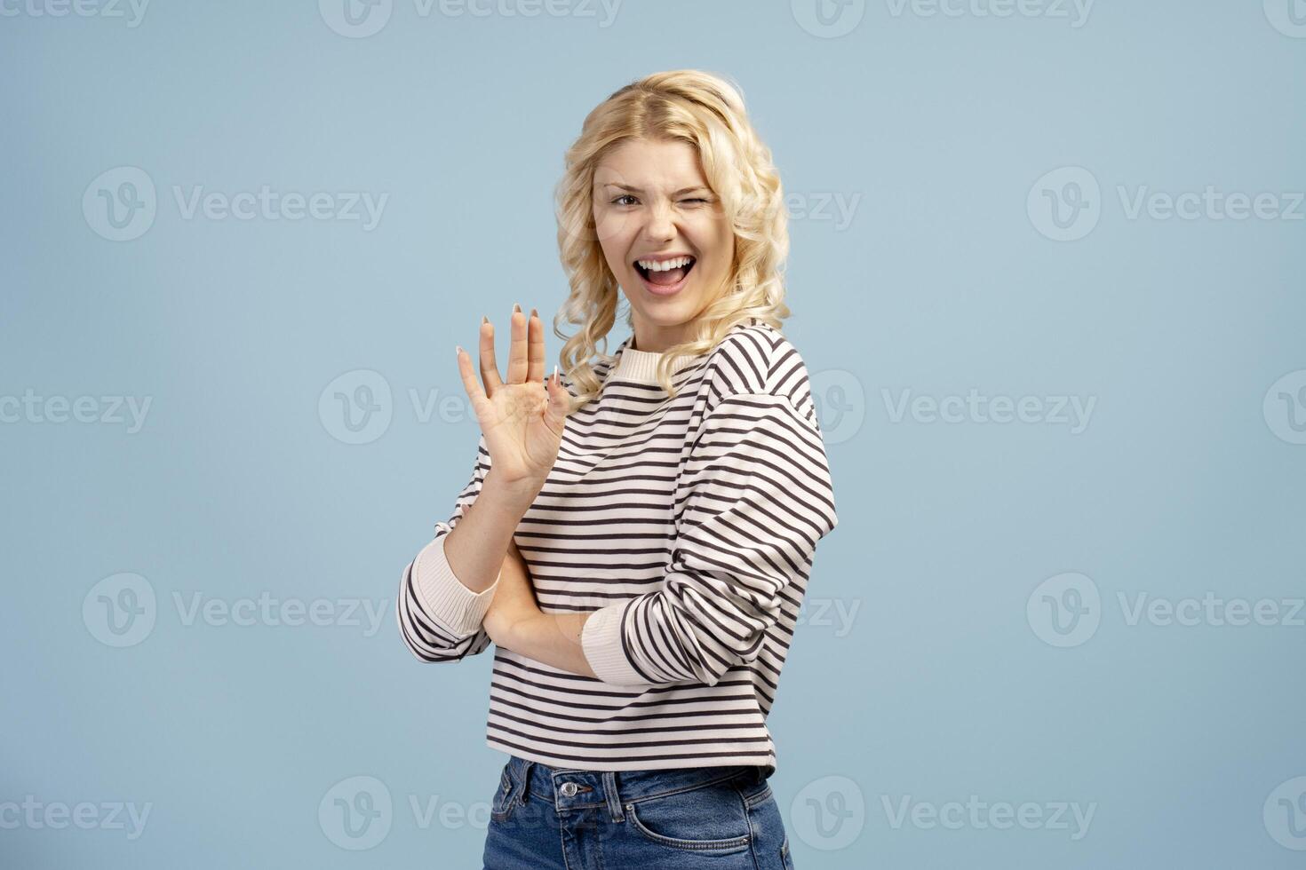 Portrait of happy, smiling blonde girl waving hand winking, posing isolated on blue background photo