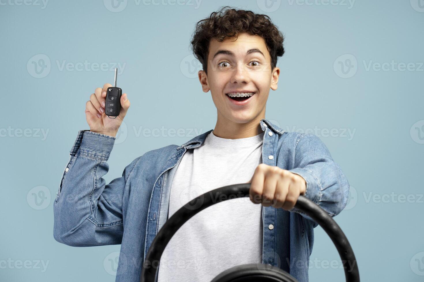 Portrait of happy, excited boy, teenager holding steering wheel and car key, on blue background photo