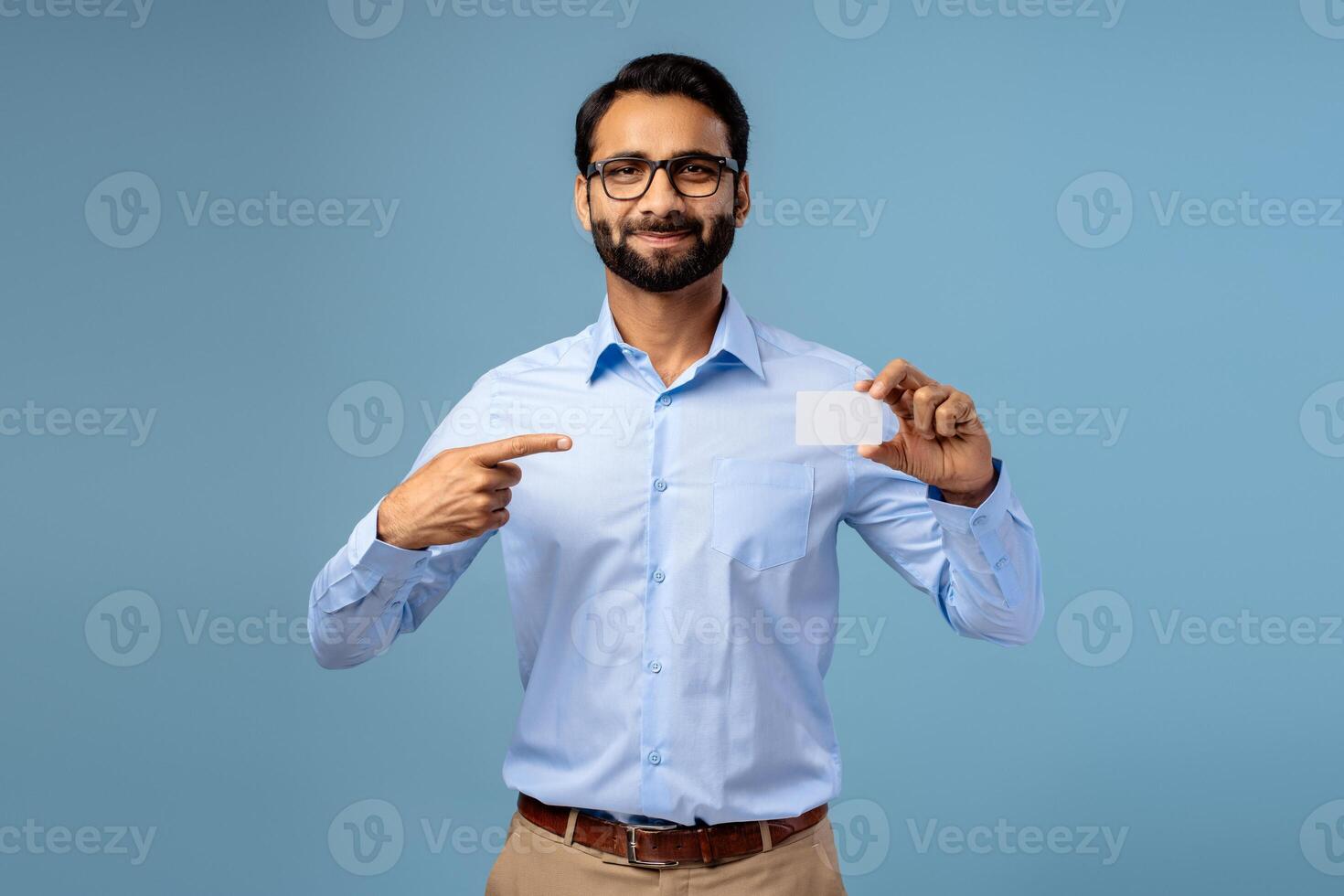 Smiling bearded handsome Indian man holding empty, white business card, pointing finger on mockup photo
