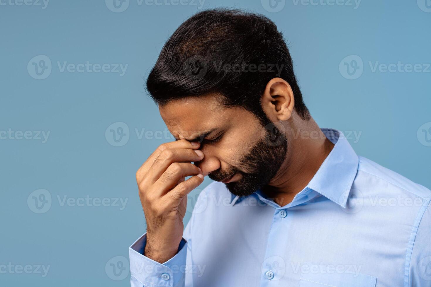 Worried Indian businessman holding head, eyes closed, showing pain and exhaustion on blue background photo