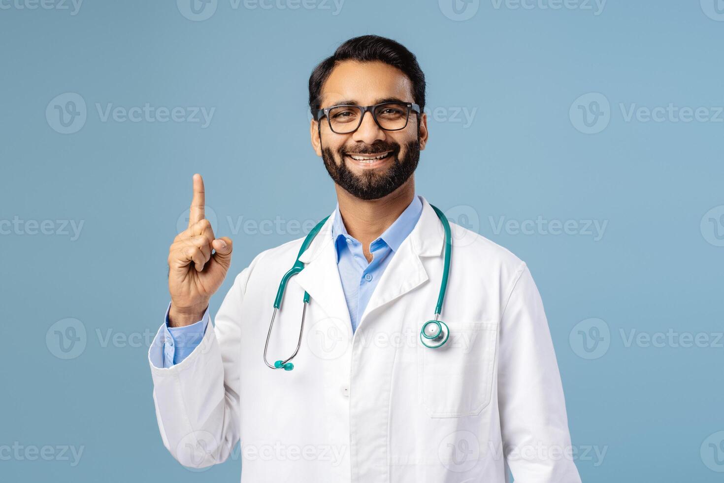 Portrait of handsome, smiling, Indian man wearing white coat, stylish eyeglasses pointing finger up photo