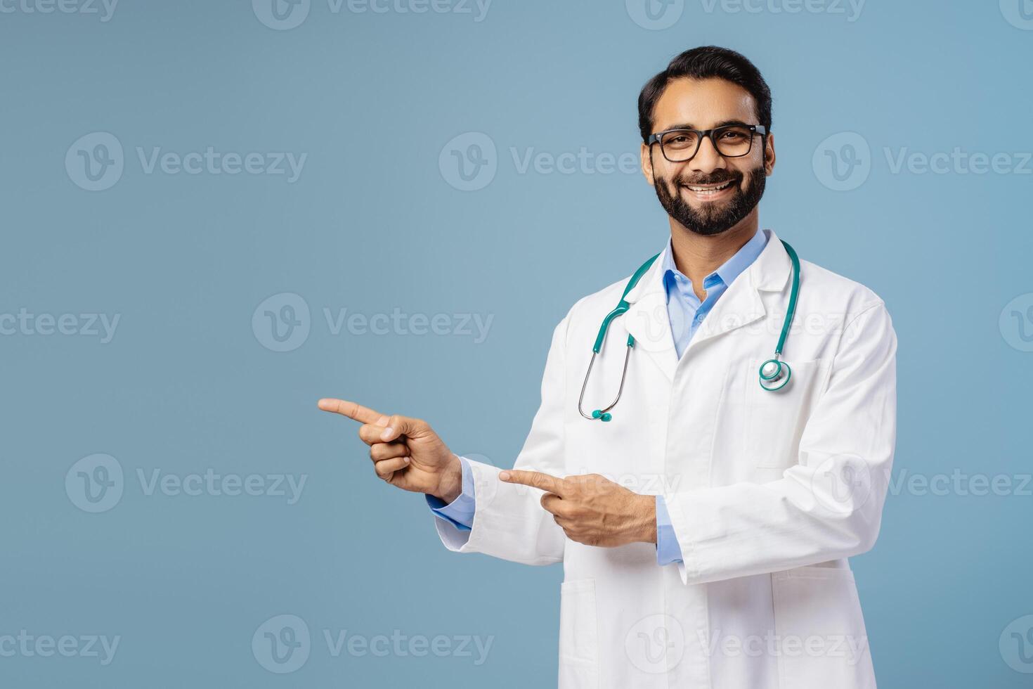 Smiling Indian man, doctor wearing white coat, stylish eyeglasses, pointing finger on copy space photo