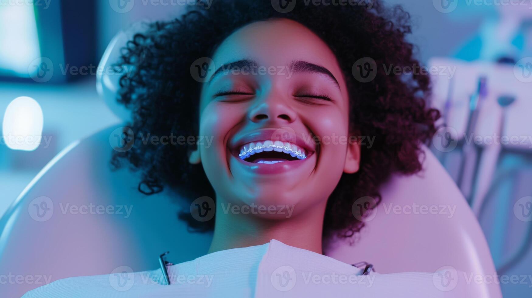 A joyful girl with curly hair showcases a wide smile, revealing her braces as she relaxes in a dental chair at an orthodontic office. photo