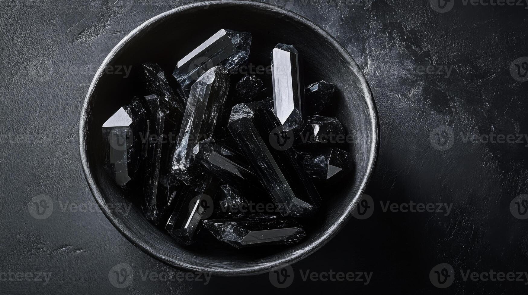 Dark black gemstones are artfully arranged in a round bowl, resting on a textured dark surface. The unique shapes and sharp angles create an intriguing visual contrast and beauty. photo