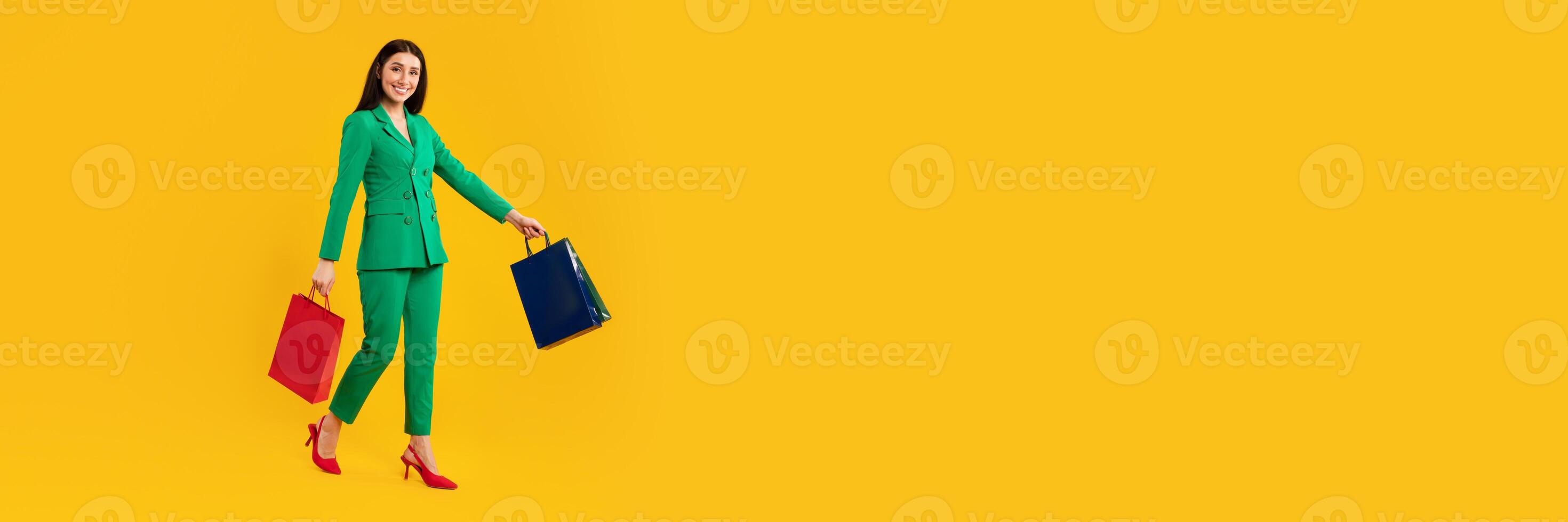 A joyful woman in a bright green suit and red heels walks while holding two shopping bags. The background is a vibrant yellow, creating a lively atmosphere perfect for Black Friday shopping. photo