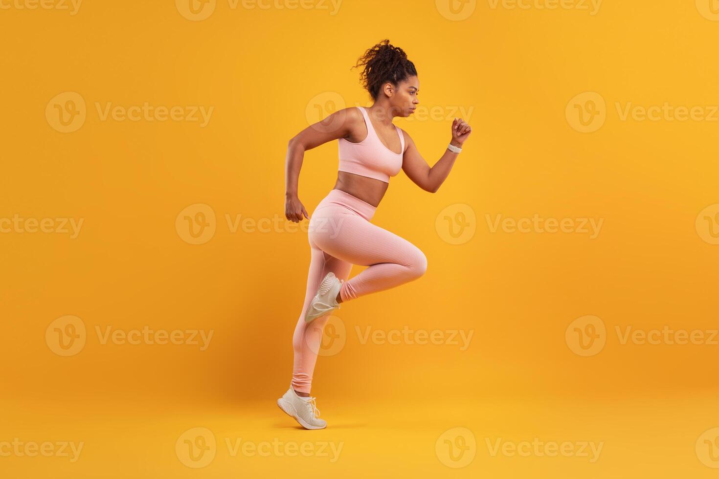 A woman in athletic wear is running energetically against a bright yellow background, showcasing her determination and focus during a workout. Her pose exudes strength and athleticism. photo