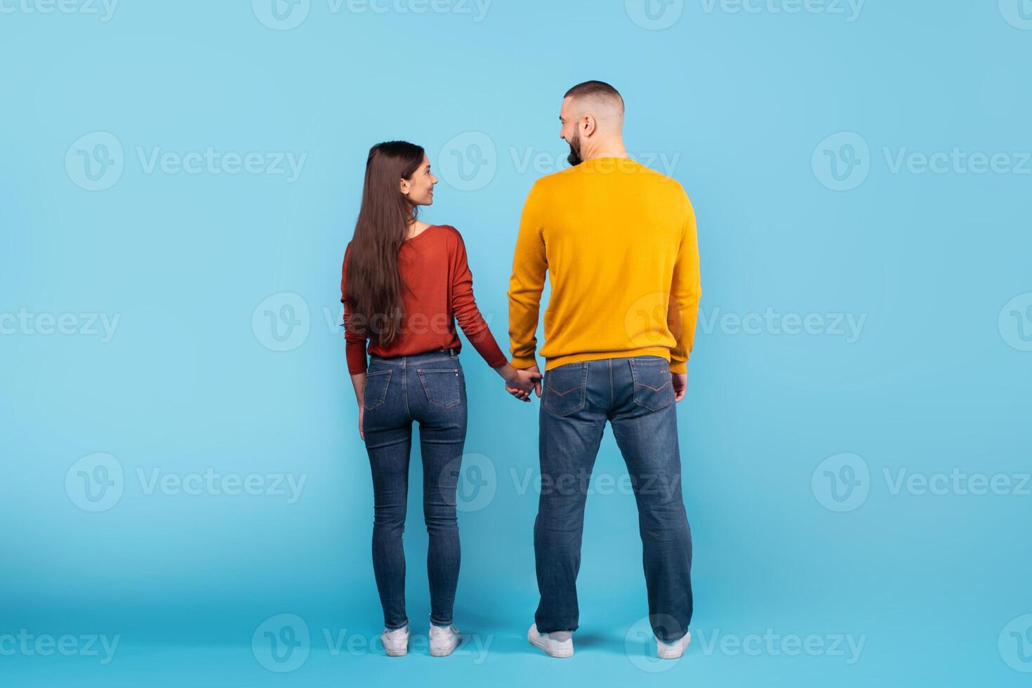 A young married couple stands together, holding hands and looking at each other affectionately. They wear casual outfits, looking relaxed and happy against a bright blue backdrop, back view photo