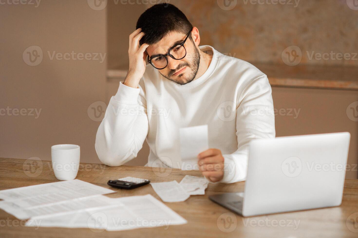 A young European man wearing eyeglasses sits at a wooden table in his home, looking puzzled as he examines a document. photo