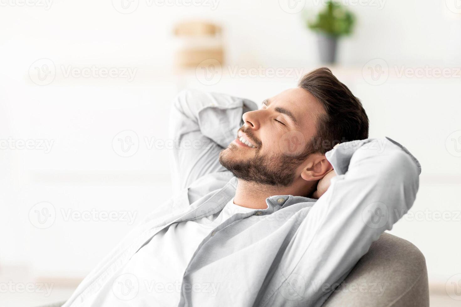 Lazy day. Happy relaxed man resting on comfortable armchair, holding hands behind head, sitting with closed eyes and enjoying silence and comfort of minimalism lifestyle, free space photo