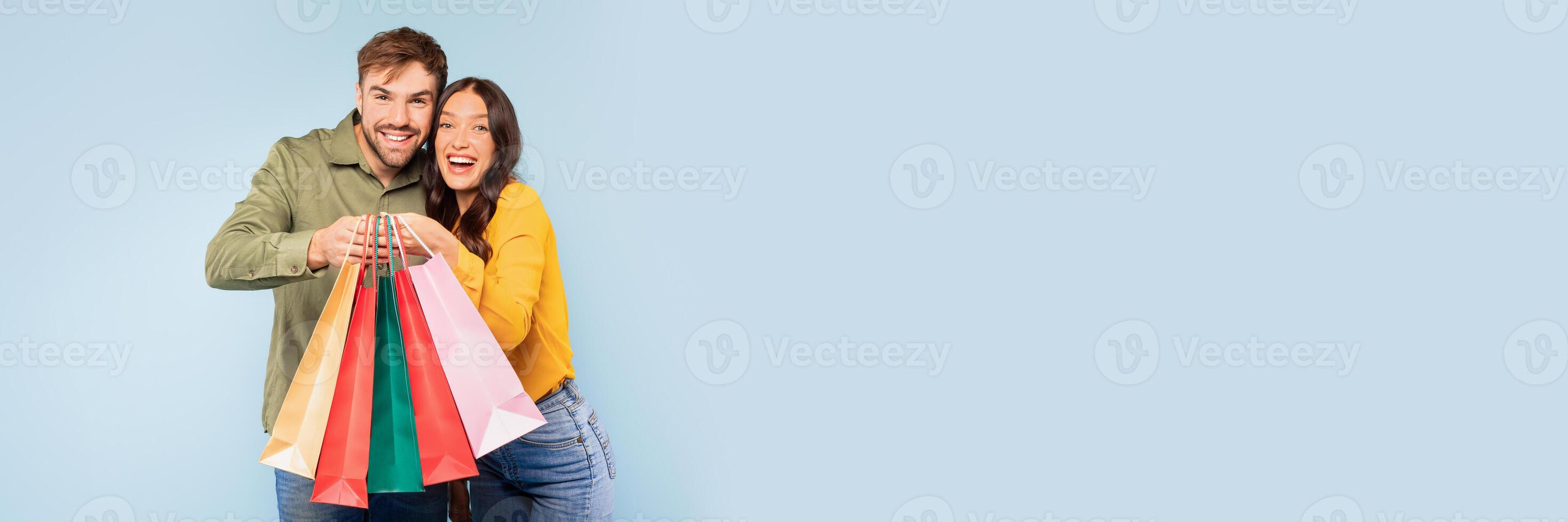 Radiant young couple holding collection of colorful shopping bags, their faces beaming with the thrill of successful shopping day, set against bright blue background, copy space photo