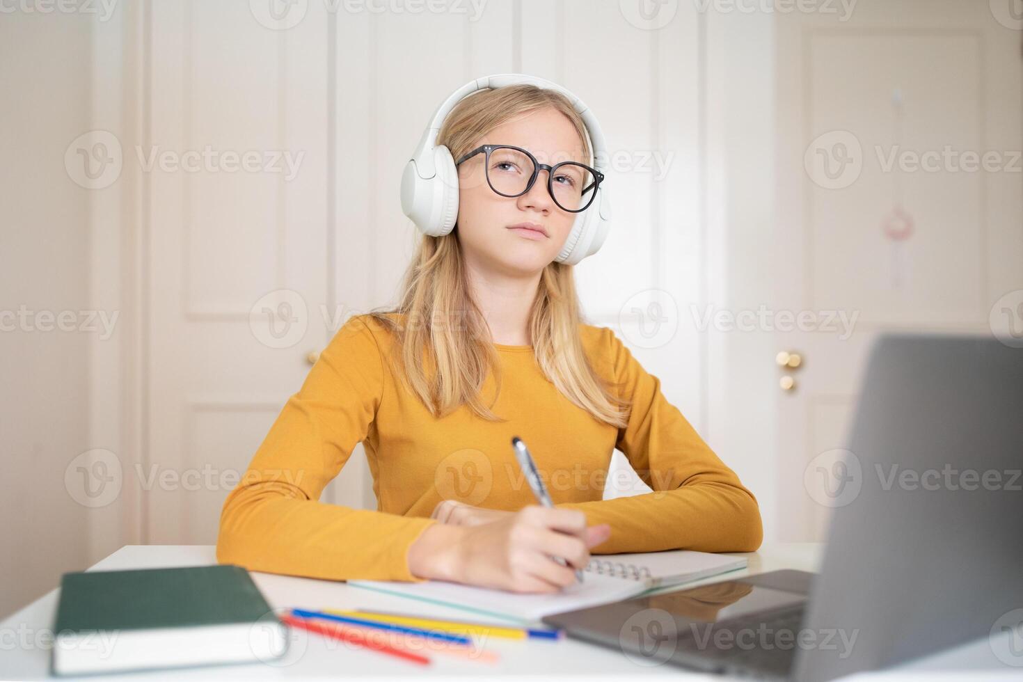 A teenage girl wearing headphones sits at a desk at home, focused on her studies. Colorful pens and a notebook are in front of her, and a laptop is open to assist with her learning. photo
