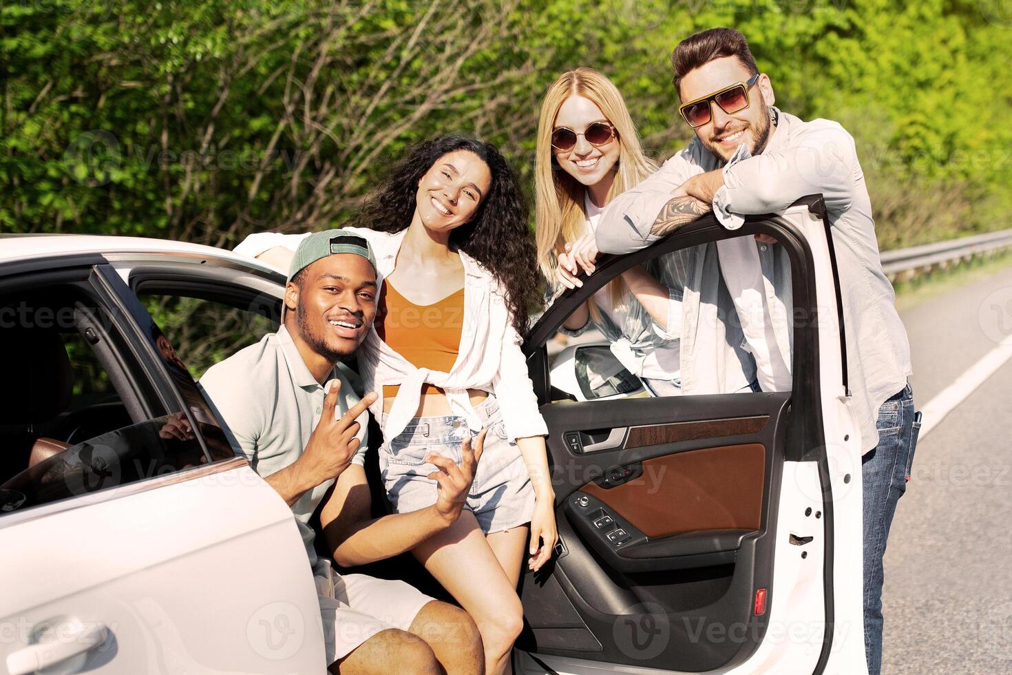 Summertime road trip, traveling with friends, joyful vacation concept. Group of diverse young people posing near their car on highway, having summer journey, enjoying fun adventure, copy space photo