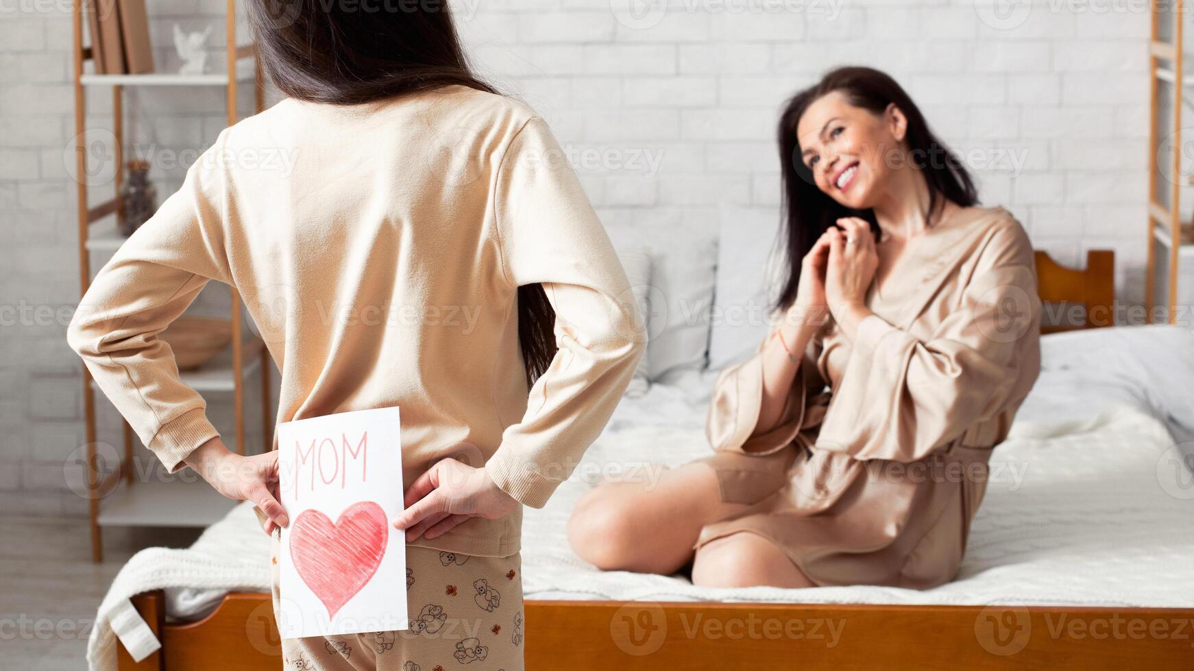 Holiday celebration. Girl hiding greeting card for mommy behind her back at home photo