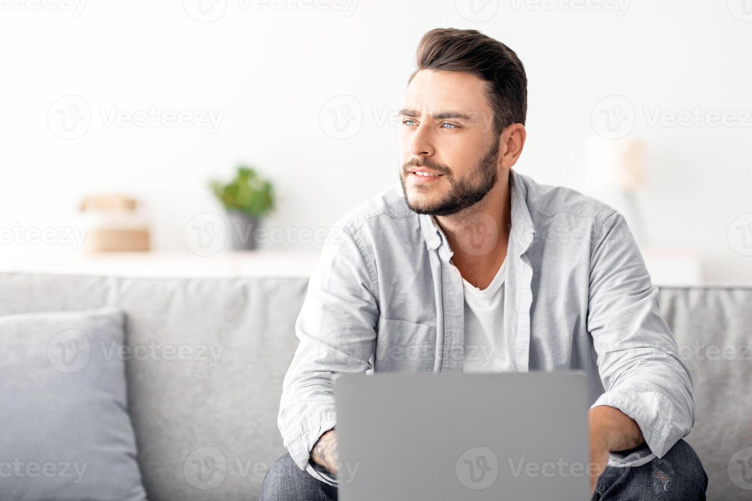 Millennial freelancer working on laptop and looking aside, thinking about successful business strategy or dreaming about holiday, sitting on sofa at home interior, free space photo