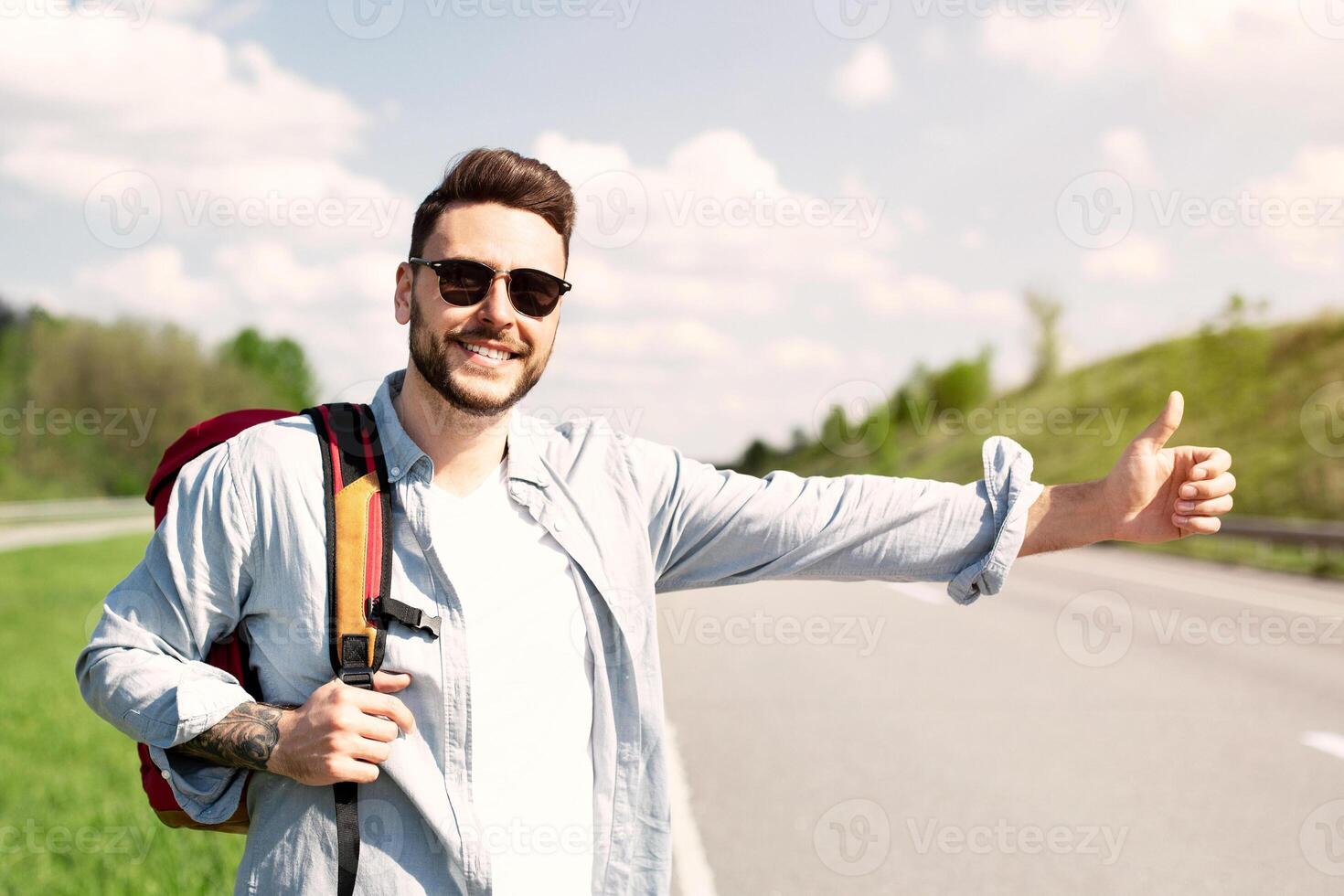 Handsome young man with backpack hitchhiking on road, showing thumb up gesture, stopping passing car. Autostop journey, summertime travel, freedom and tourism concept photo