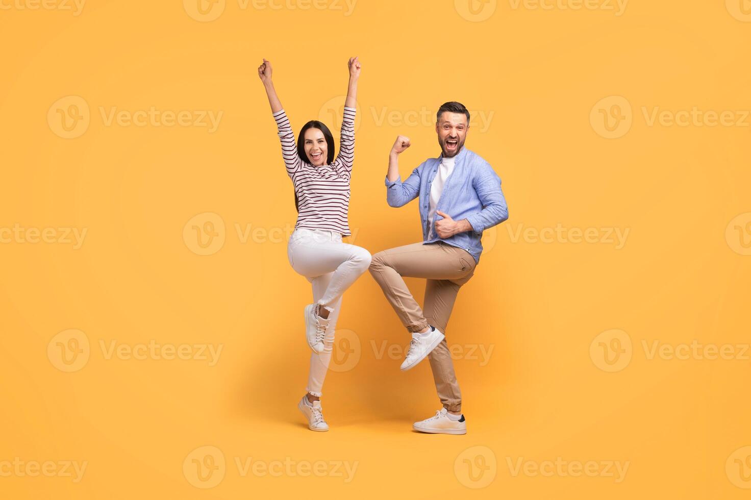 A happy couple stands on a bright yellow backdrop, both wearing casual attire. They express joy, raising their arms and smiling at one another, celebrating their love and togetherness. photo
