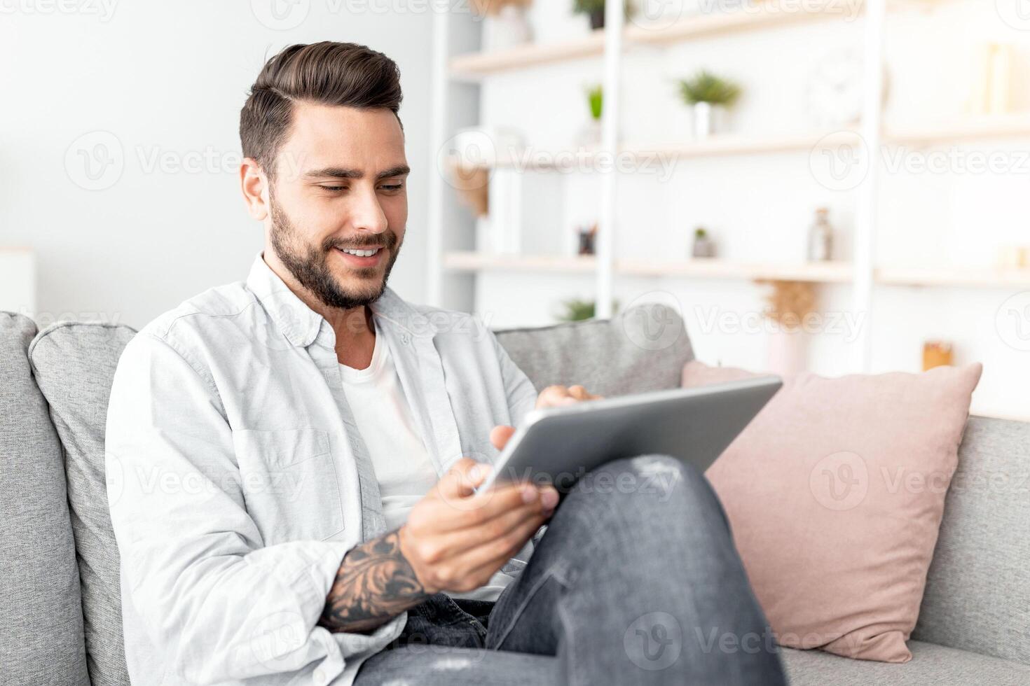 Relaxed millennial man resting on sofa with digital tablet, free space. Positive guy browsing social networks or shopping online while sitting in living room interior photo