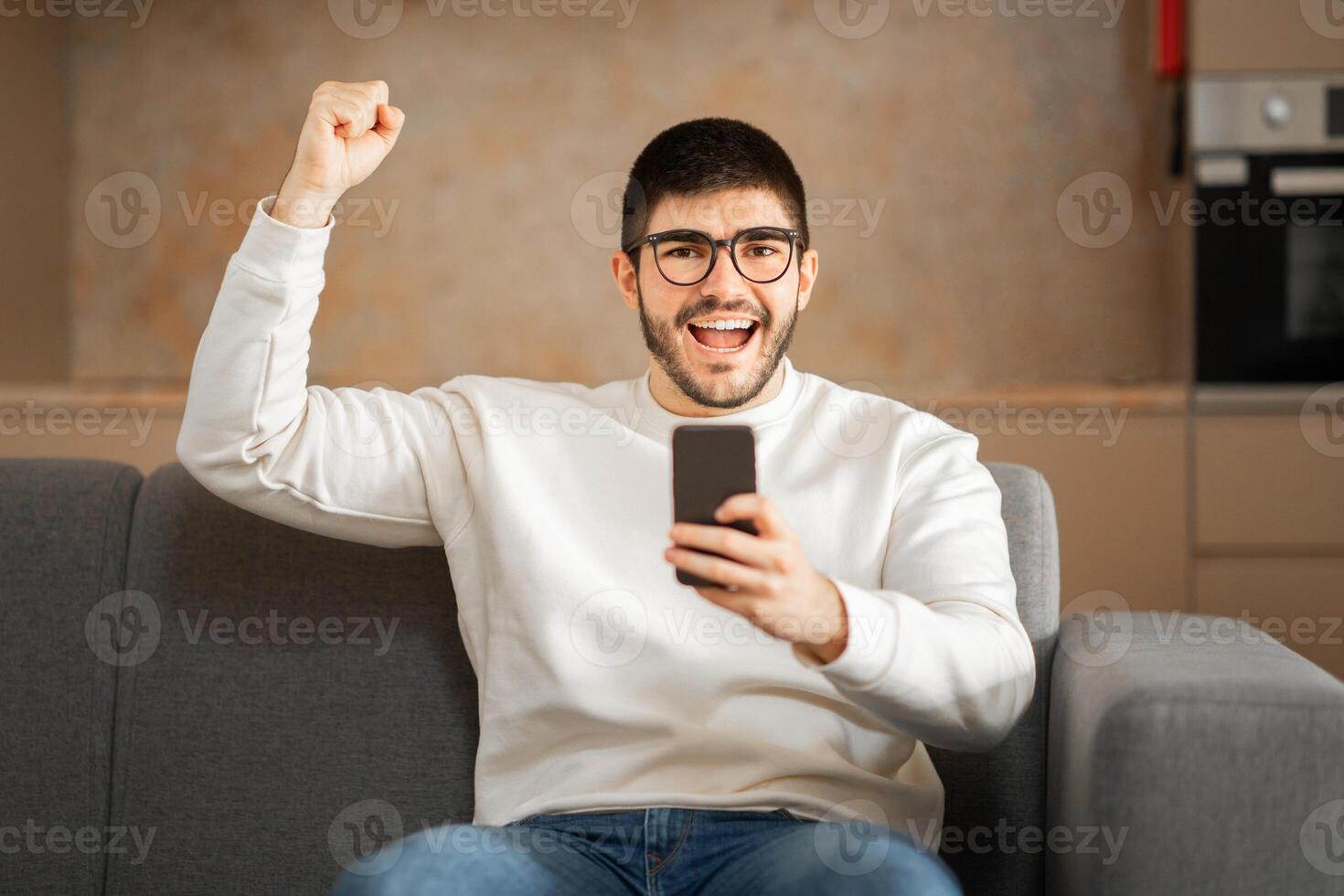A young Caucasian man wearing eyeglasses expresses excitement on a couch in his home. He raises his fist in joy while looking at his smartphone, enjoying a personal moment. photo