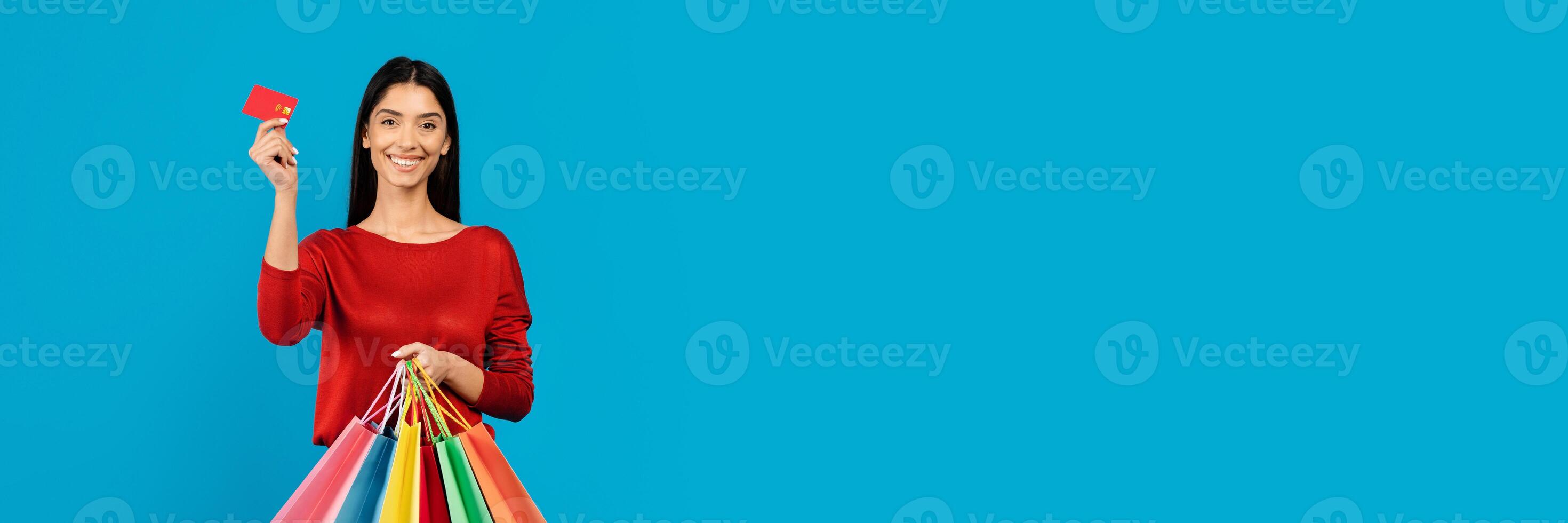 Young happy woman holding credit card in one hand and bright shopper bags in the other, happy lady ready for shopping spree, standing on blue studio background, copy space photo