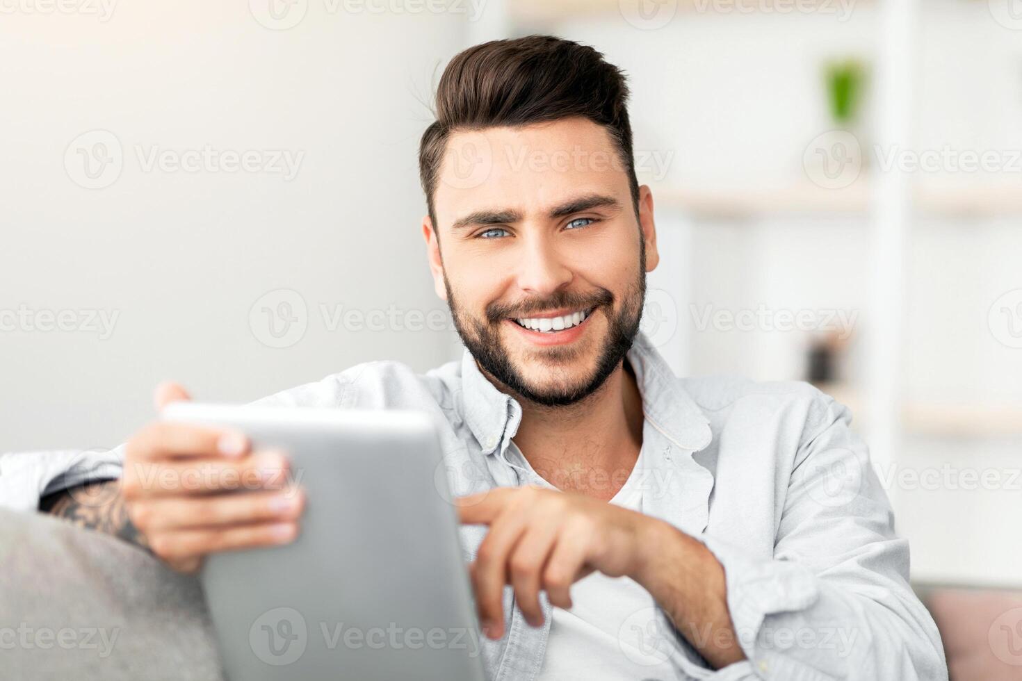Handsome young man holding digital tablet and smiling at camera, browsing internet or checking social networks, sitting on couch at home, free space. Happy guy using modern technologies for leisure photo