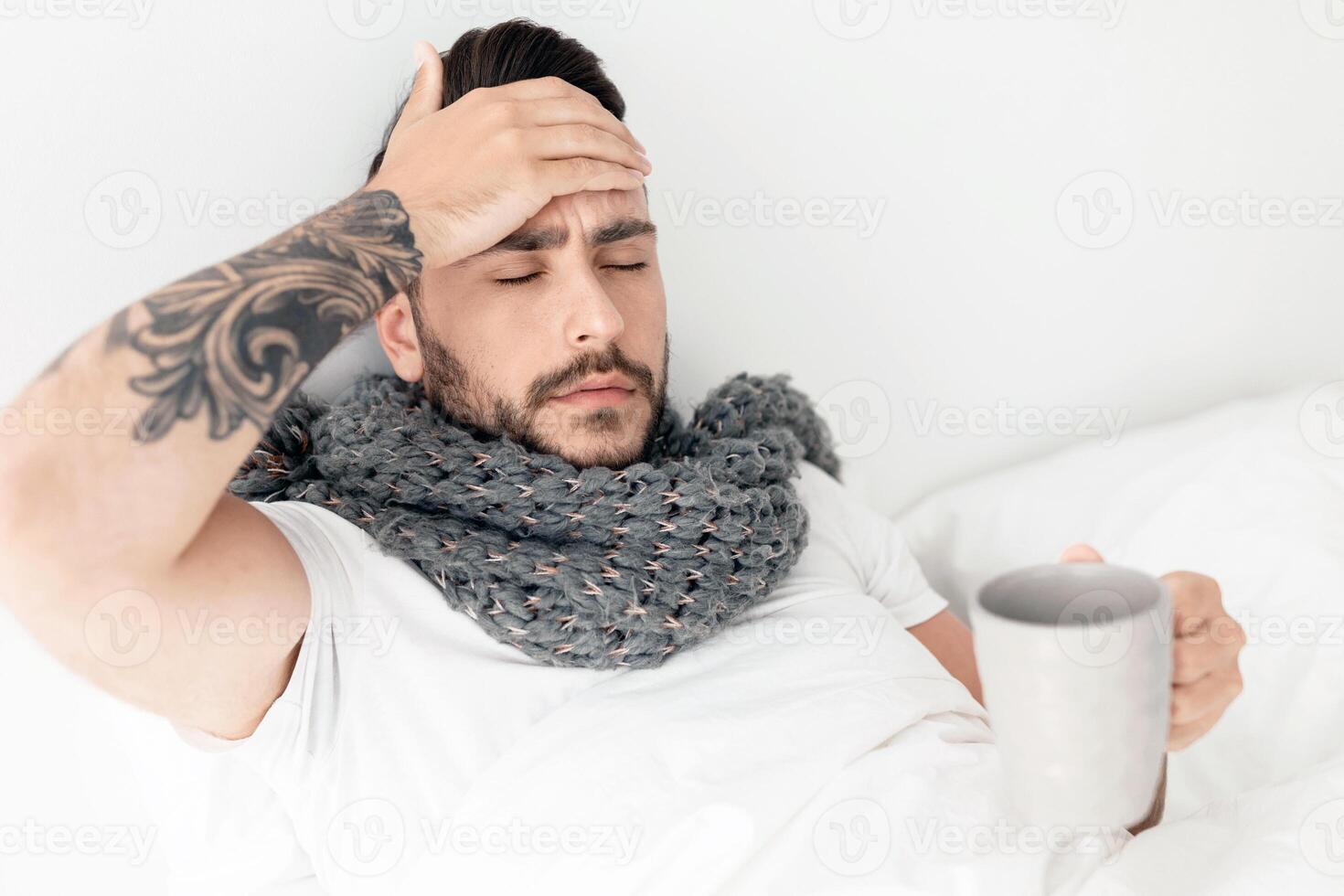 Feeling bad. Sick guy having fever, drinking hot healing tea and touching his forehead, lying in bed, empty space. Influenza symptom and coronavirus concept photo