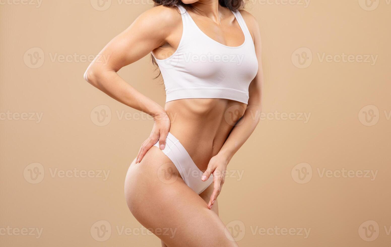 A young Caucasian woman confidently poses in white underwear, showcasing her figure against a neutral beige backdrop. The focus is on beauty and body confidence. photo