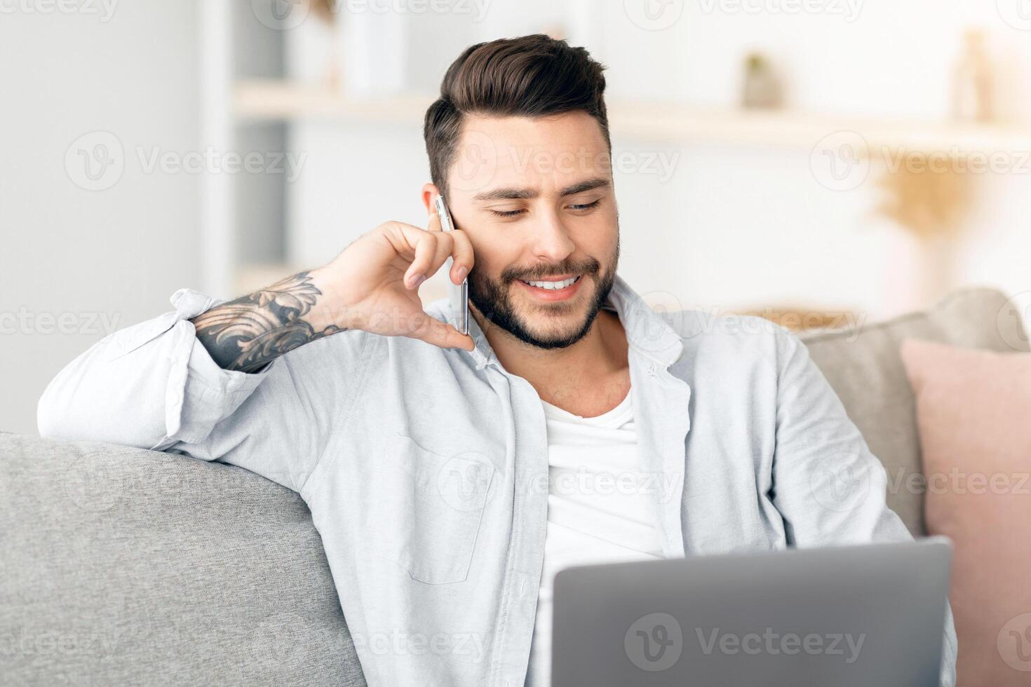 Always in touch. Young freelancer man talking on smartphone and using laptop while working remotely from home, sitting on sofa in living room. Freelance career, distant work and mobile communication photo