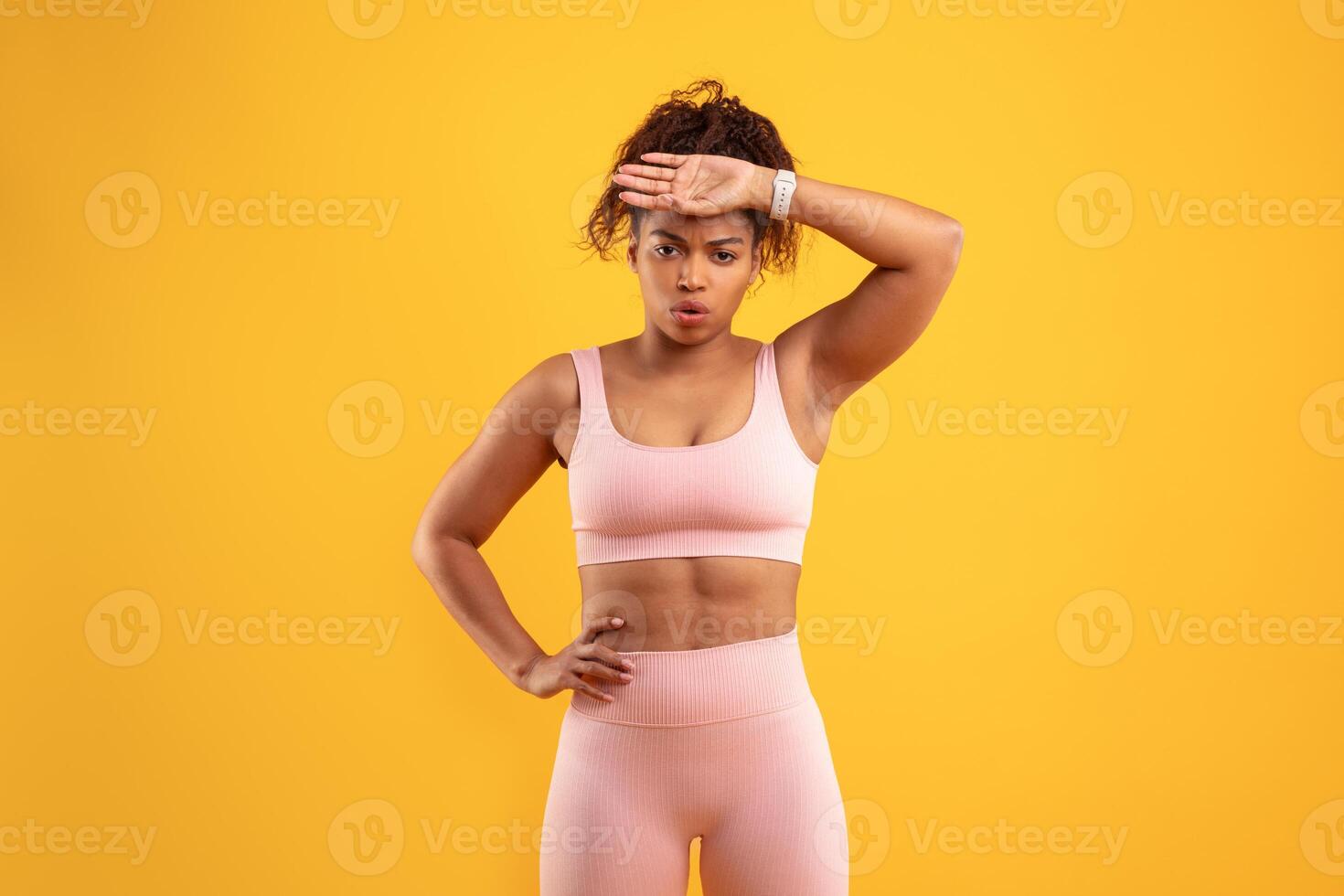 A determined woman wearing workout attire stands with one hand on her hip and the other on her forehead. Her expression shows focus and effort in a fitness-related activity. photo