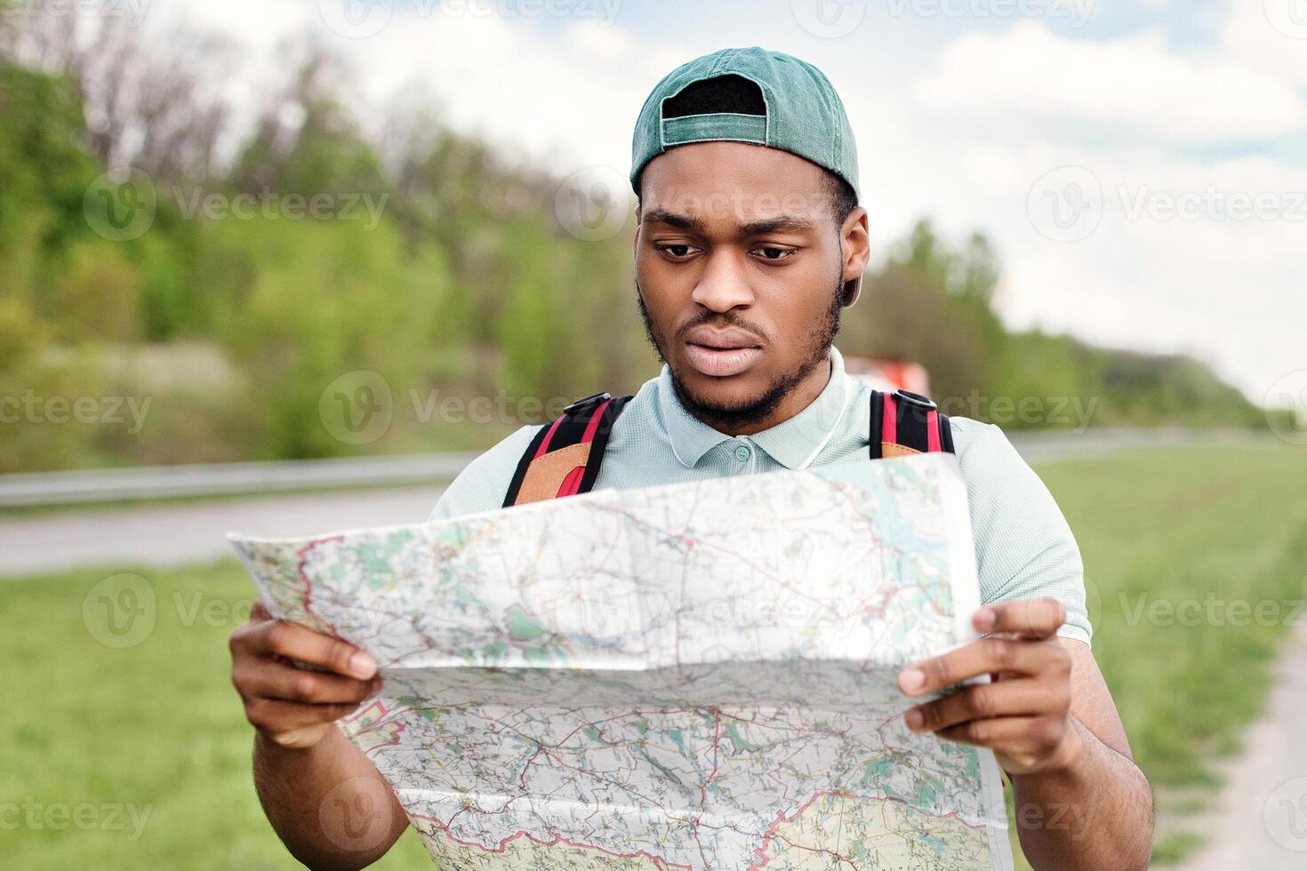 Unhappy young black man hitchhiking on road, looking at map, feeling lost, traveling alone by autostop. Millennial African American guy standing on highway, cannot find his tourist destination photo