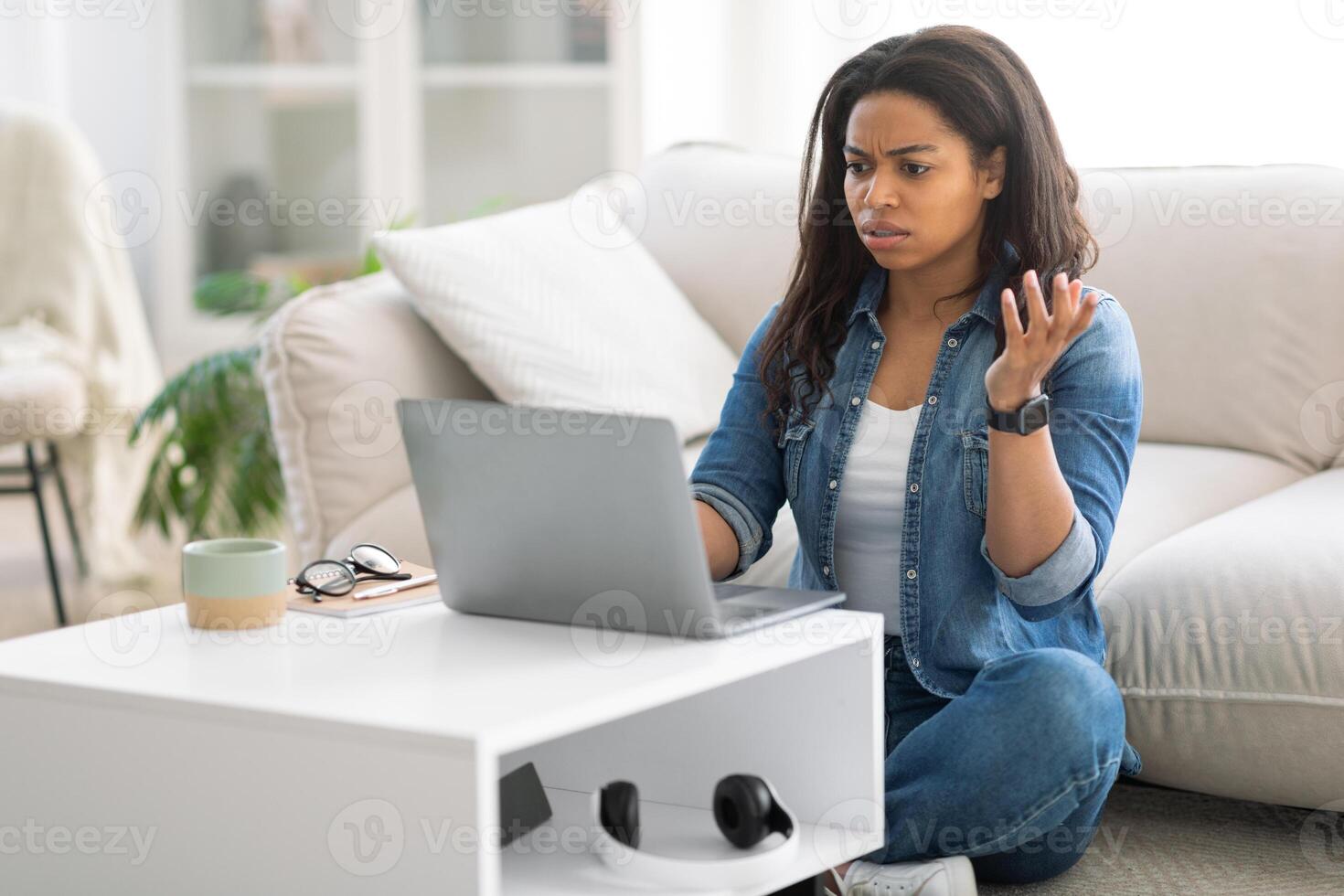 A black woman sits on the floor of her cozy living room, looking exasperated while using her laptop. She is dressed casually and appears to be dealing with a frustrating issue as she gestures. photo