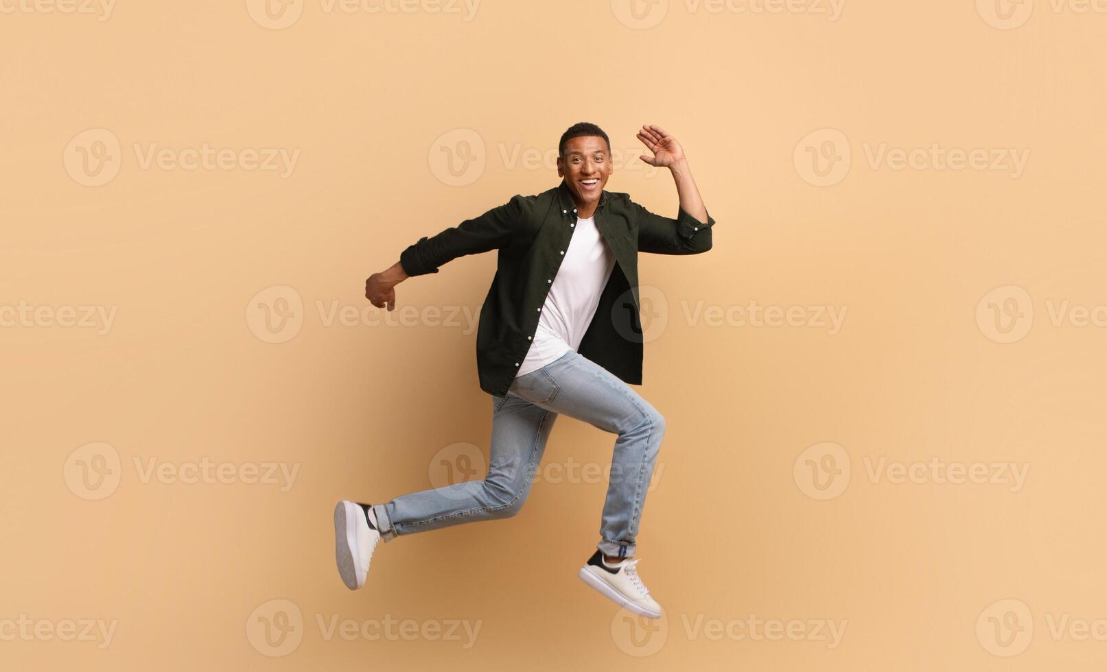 A joyful African American man is leaping in a dynamic pose. He wears casual attire with jeans and a shirt, showcasing an upbeat lifestyle while posed against a soft, beige backdrop. photo