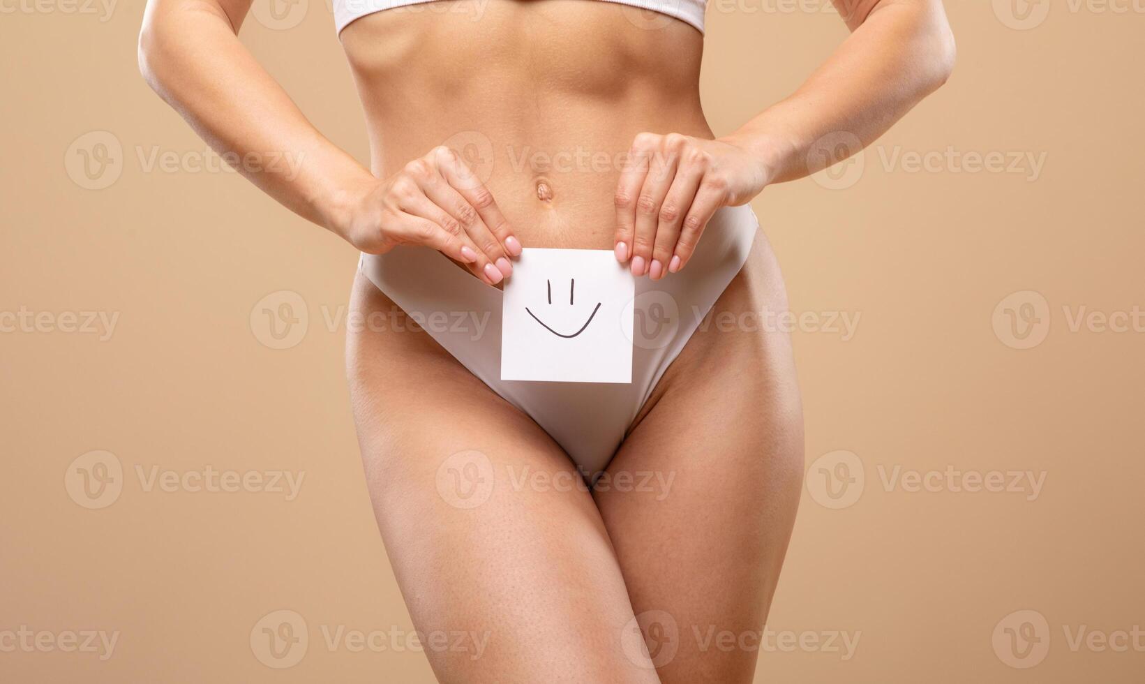 A young Caucasian woman poses gracefully in elegant lingerie, playfully holding a card with a smiley face. The neutral background enhances her beauty and confidence. photo