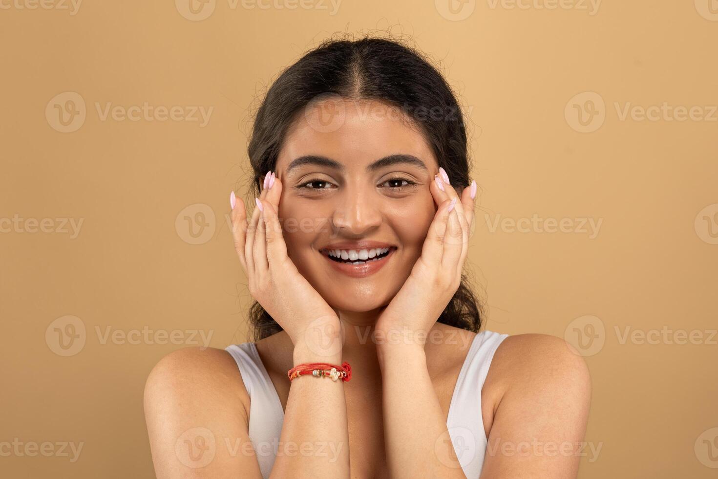 A young Indian woman with a joyful expression pampering her skin. She is using skincare products, highlighting self-care and beauty. The warm background enhances her glowing complexion. photo