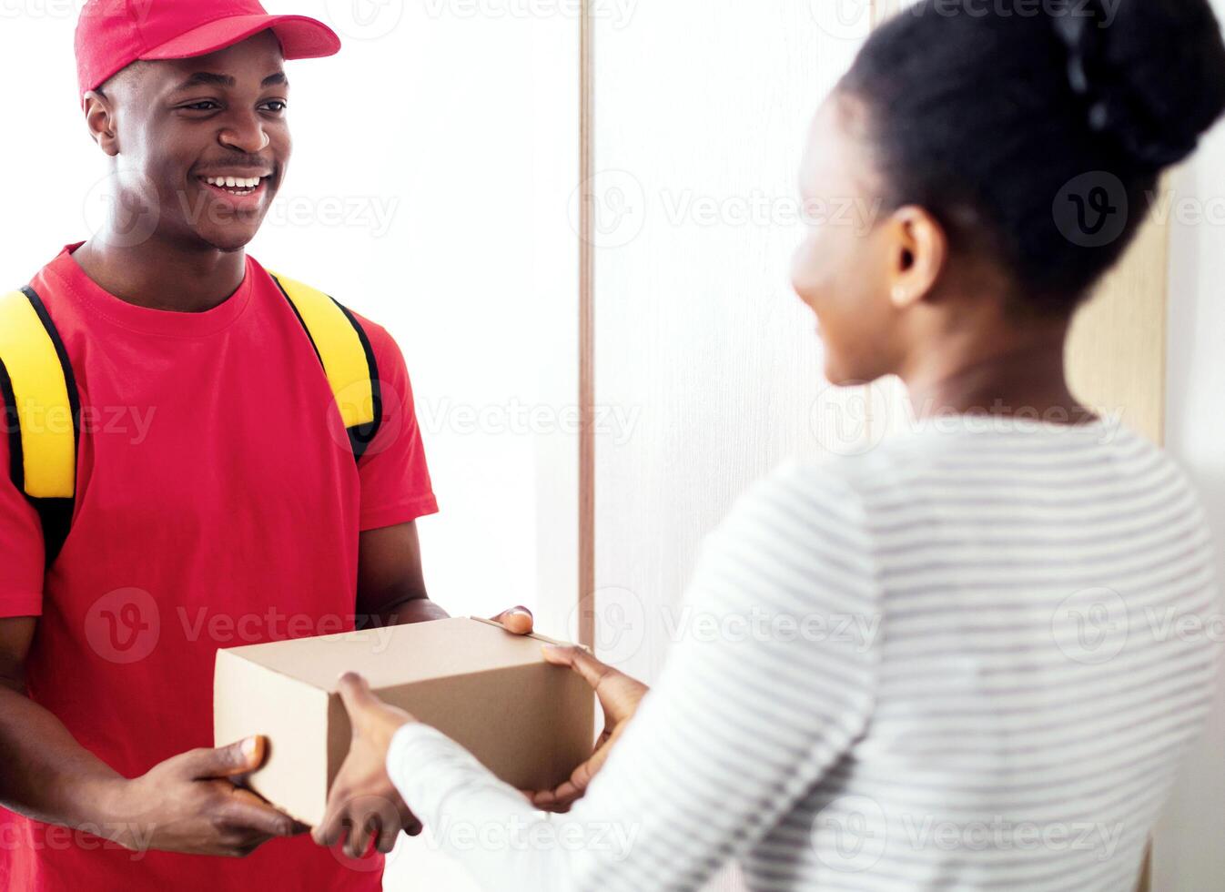 Cheerful Black Courier Man Delivering Cardboard Parcel Box To Woman Standing At Her Door Indoors. Fast Delivery Service And Postal Shipping Concept. Selective Focus photo
