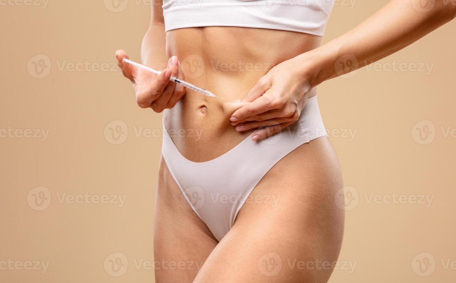 A young European woman in white lingerie is applying a skin treatment to her abdomen. She stands in a minimalistic indoor setting, focusing on her beauty routine and self-care. photo