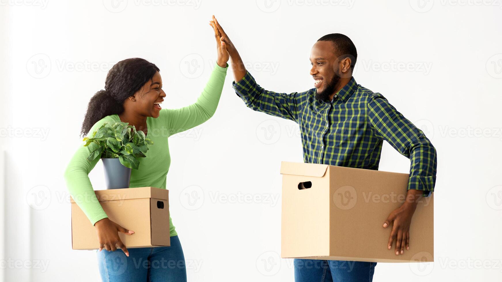 Happy Moving Day. Joyful Black Spouses Holding Cardboard Boxes And Giving High Five To Each Other, Cheerful African American Couple Celebrating Relocation To Their Own Apartment, Copy Space photo