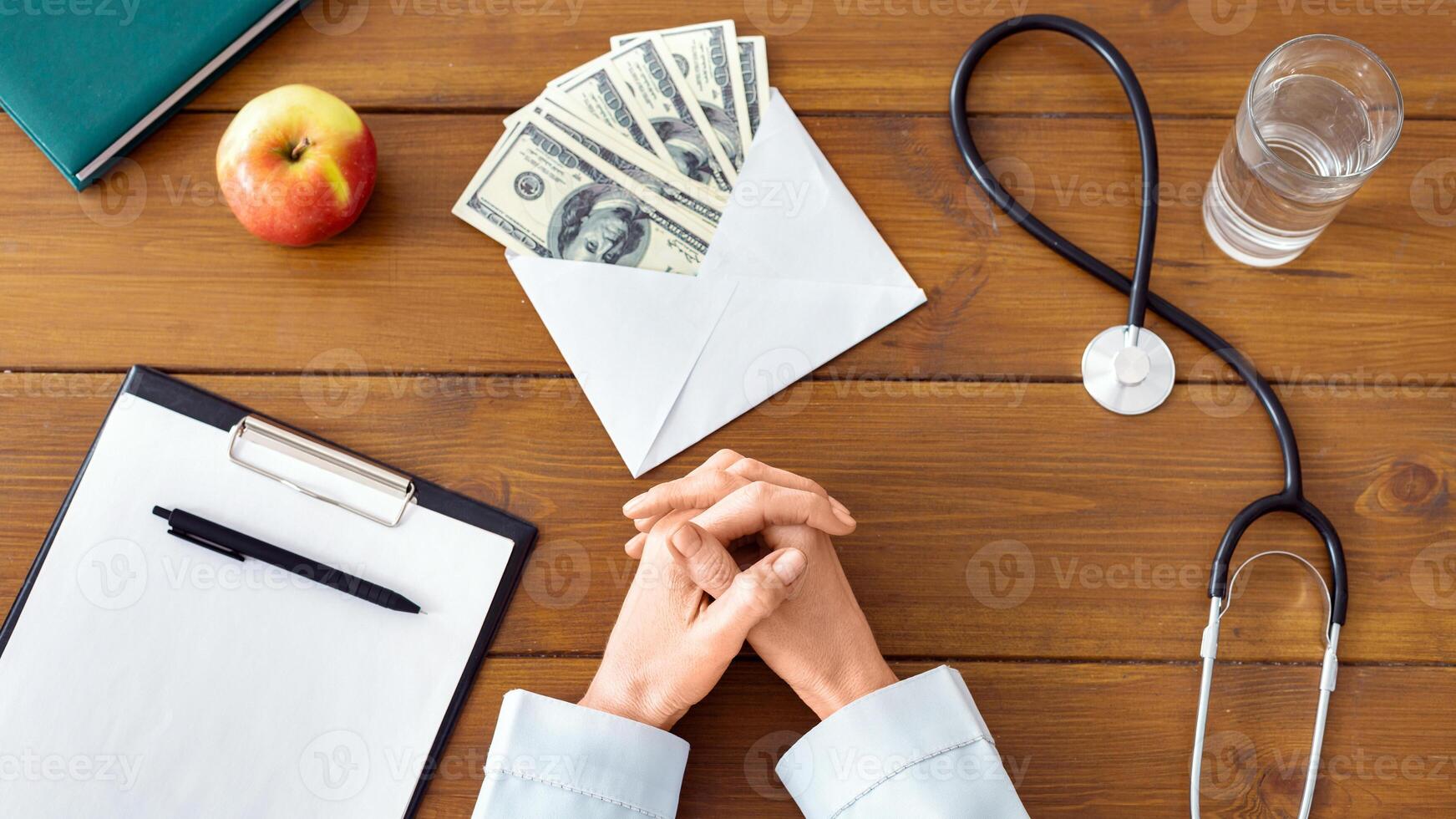 Corruption concept. Crossed female doctor hands on wooden table with envelope with money, cropped, top view photo