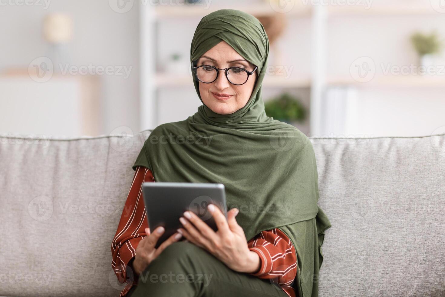 Senior Muslim Female Using Digital Tablet Reading Book Online Sitting On Couch At Home. Modern Middle-Eastern Housewife In Hijab And Glasses Watching Movie, Browsing Internet Via Computer photo