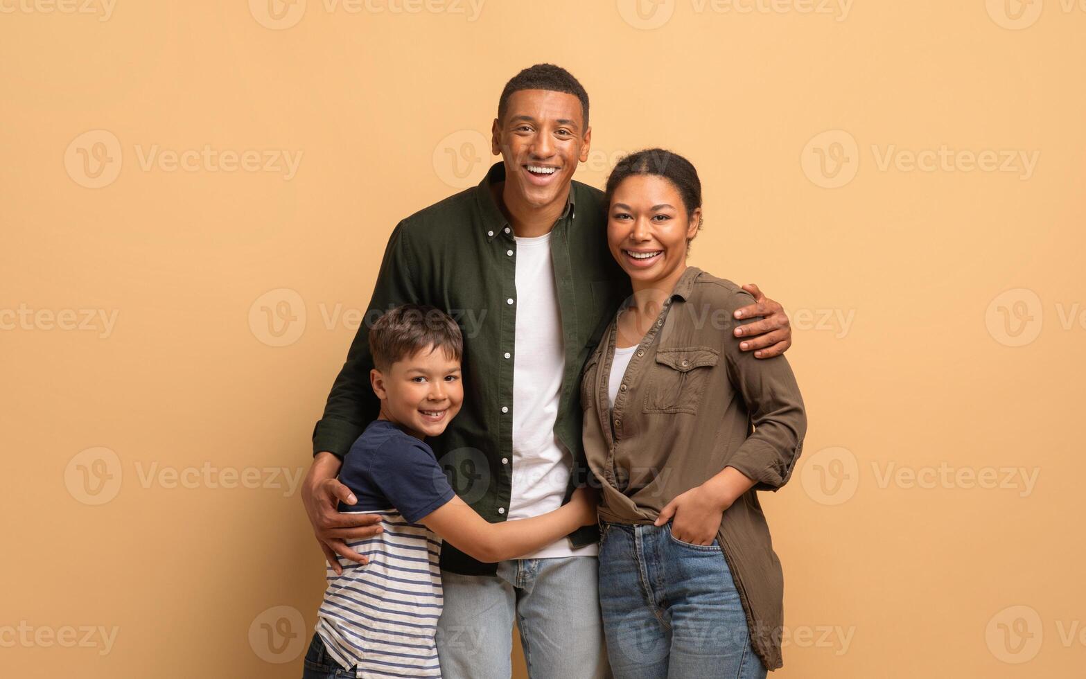 A loving African American family shares a warm embrace, with a father, mother, and son smiling joyfully against a neutral background. Their bond reflects happiness and togetherness. photo