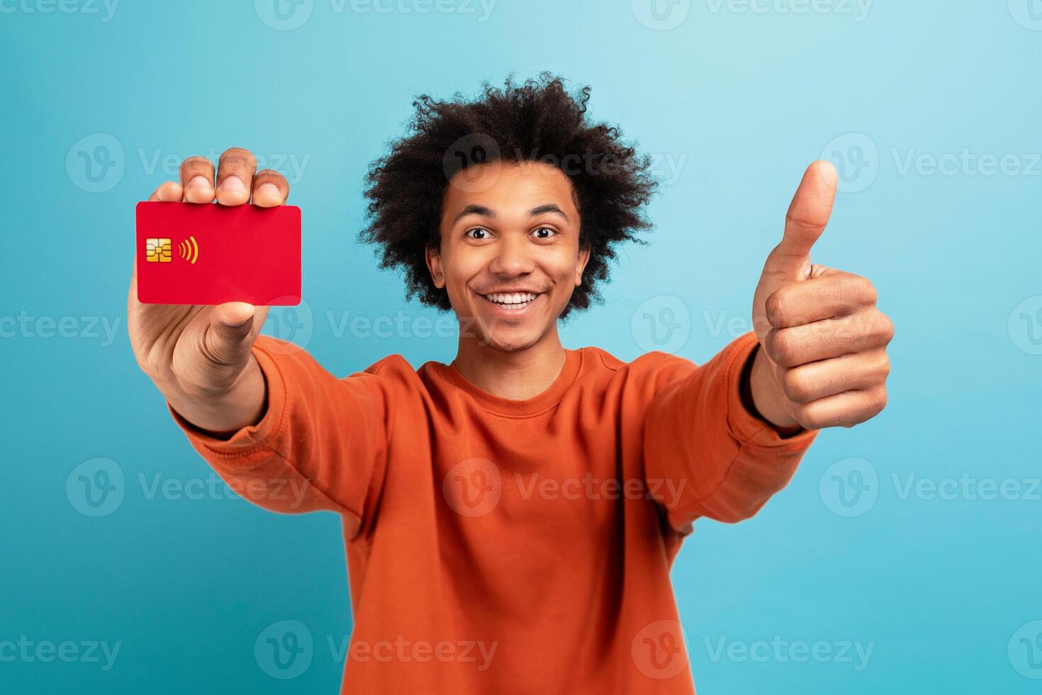 A young African American man smiles while holding a red card and giving a thumbs up. He has curly hair and wears an orange sweater, standing in front of a light blue backdrop. photo